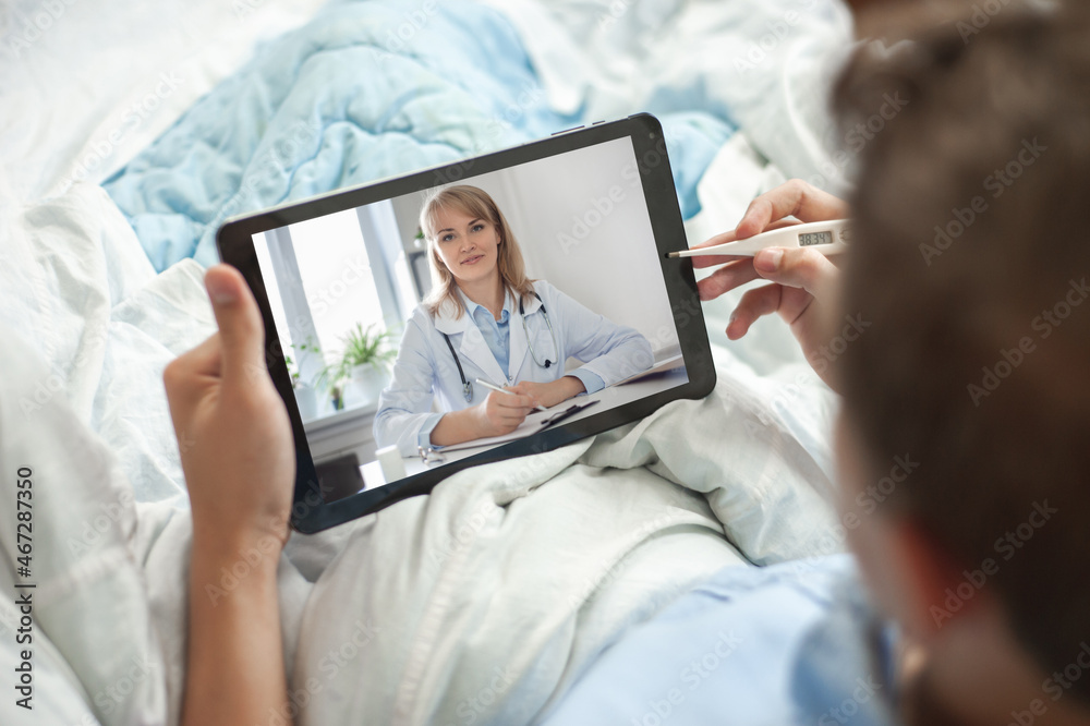 Telemedicine and online consultation concept. A young man or teenager sits in bed with an electronic thermometer and a tablet in his hands. The young man has video chat with a doctor.