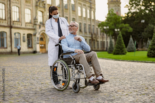 Full length shot of senior man handicapped patient in wheelchair smiling at young nurse in protective mask helping him outdoors