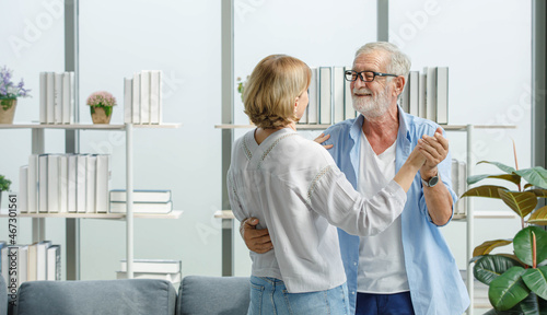 Caucasian old senior elderly happy grandparents lover married couple standing smiling look at each other face listening to slow waltz music dancing together in living room at home in front bookshelf