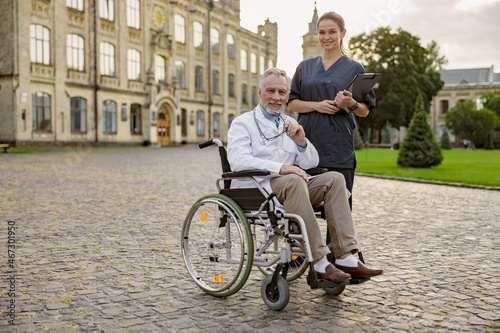 Full length shot of senior handicapped male doctor in wheelchair wearing lab coat holding glasses while posing together with young nurse assisting him outdoors