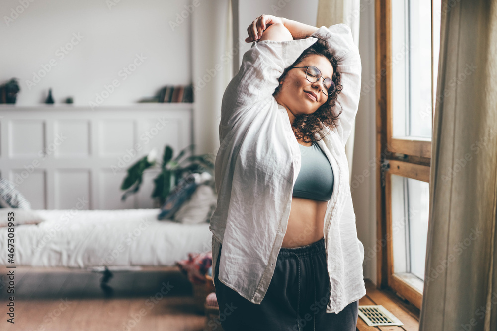 Fototapeta premium Curly haired overweight young woman in blue top and shorts with satisfaction on face accepts curvy body shape in stylish bedroom.