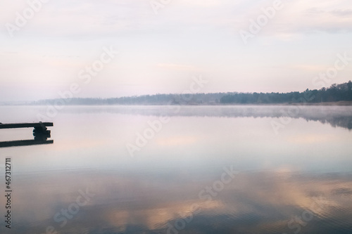 General plan of the pier for fishermen, which is located on a misty lake. The mist on the water creates the feeling of a magical effect. Clouds bounce off the surface. Place for text