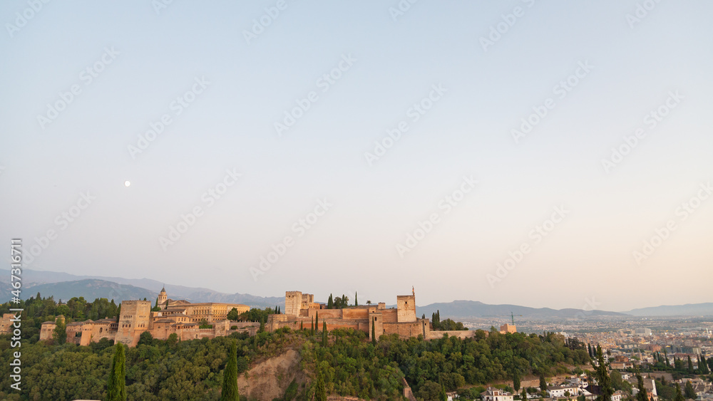 Aerial view of the famous Alhambra at sunset, Granada, Andalusia, Spain. White moon on the left. Pale blue sky on the background and view of the city on the right.