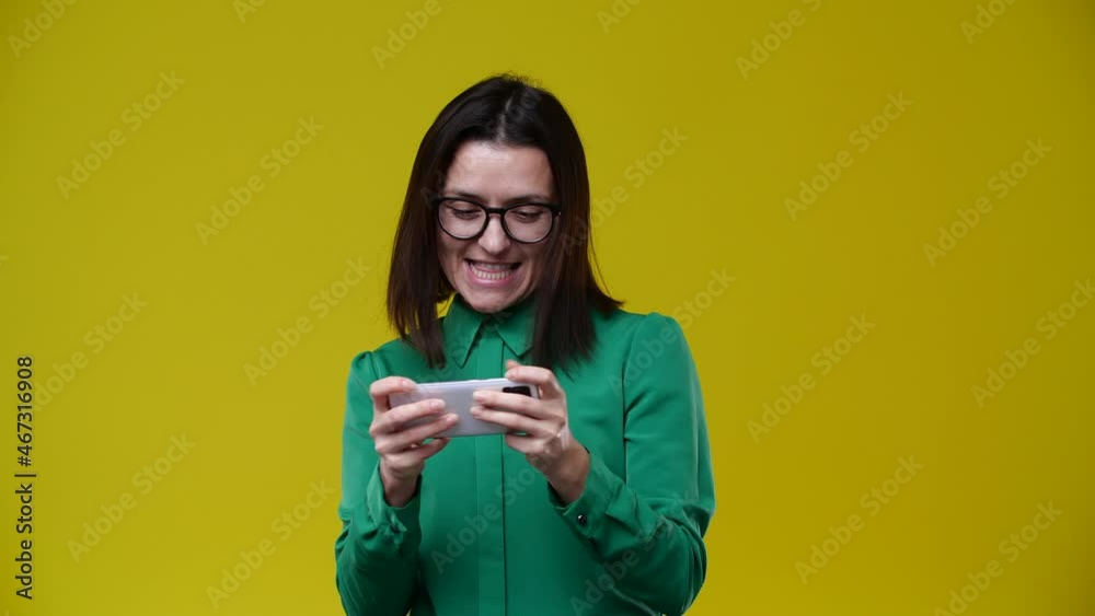 the girl plays games on the phone, races, and wins, she is brunette, has short hair, wears glasses, is wearing a green shirt, the background is yellow the girl plays games on the phone, races, and wins, she is brunette, has short hair, wears glasses, is wearing a green shirt, the background is yellow