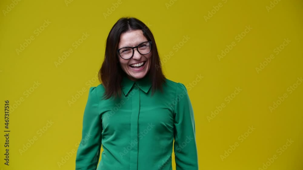 the woman waves her fists and she is very cheerful, the smile does not leave her face, she has dark hair, wears glasses, is dressed in a green shirt, the background is yellow the woman waves her fists and she is very cheerful, the smile does not leave her face, she has dark hair, wears glasses, is dressed in a green shirt, the background is yellow