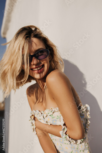 Happy caucasian lady laughing looking to side against background of white wall. Blonde in translucent glasses holds her sundress on chest with bare shoulders. Concept of recreation, healthy lifestyle