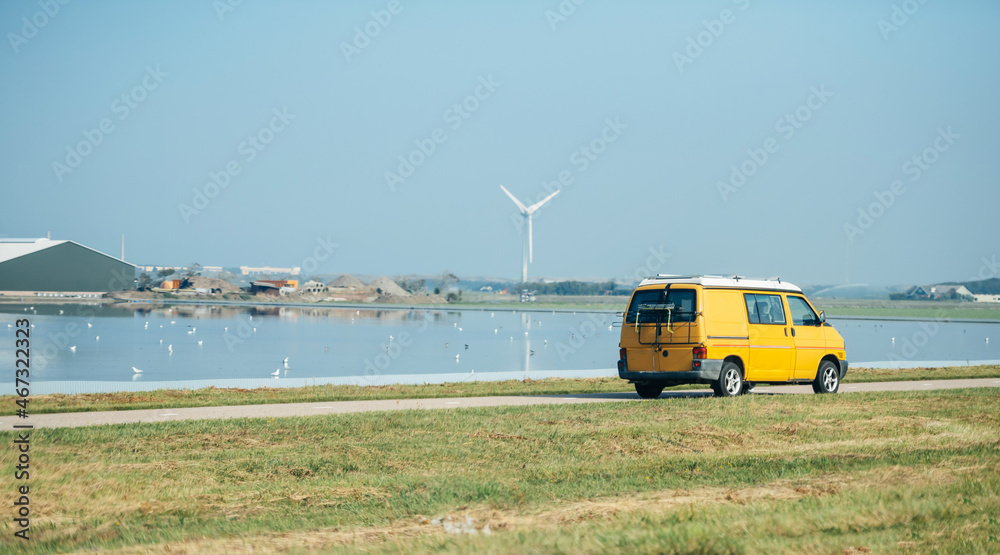 Yellow travel bus driving fast near water canal in Netherlands on the coast with tall wind turbine and multiple birds on the calm waters Yellow travel bus driving fast near water canal in Netherlands on the coast with tall wind turbine and multiple birds on the calm waters