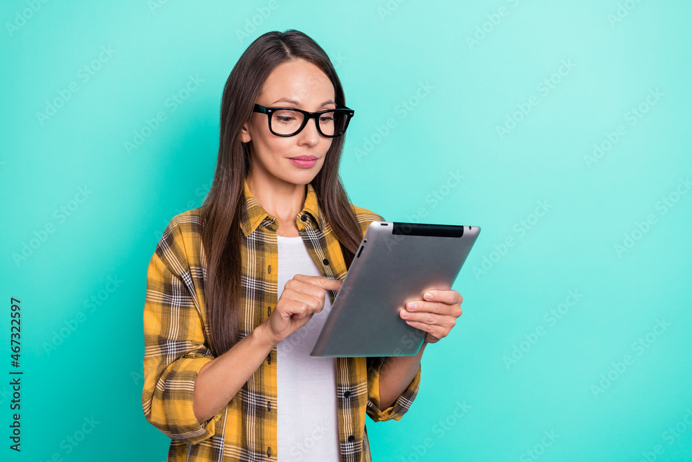 Photo of bossy young brunette lady hold tablet wear eyewear yellow shirt isolated on turquoise color background Photo of bossy young brunette lady hold tablet wear eyewear yellow shirt isolated on turquoise color background
