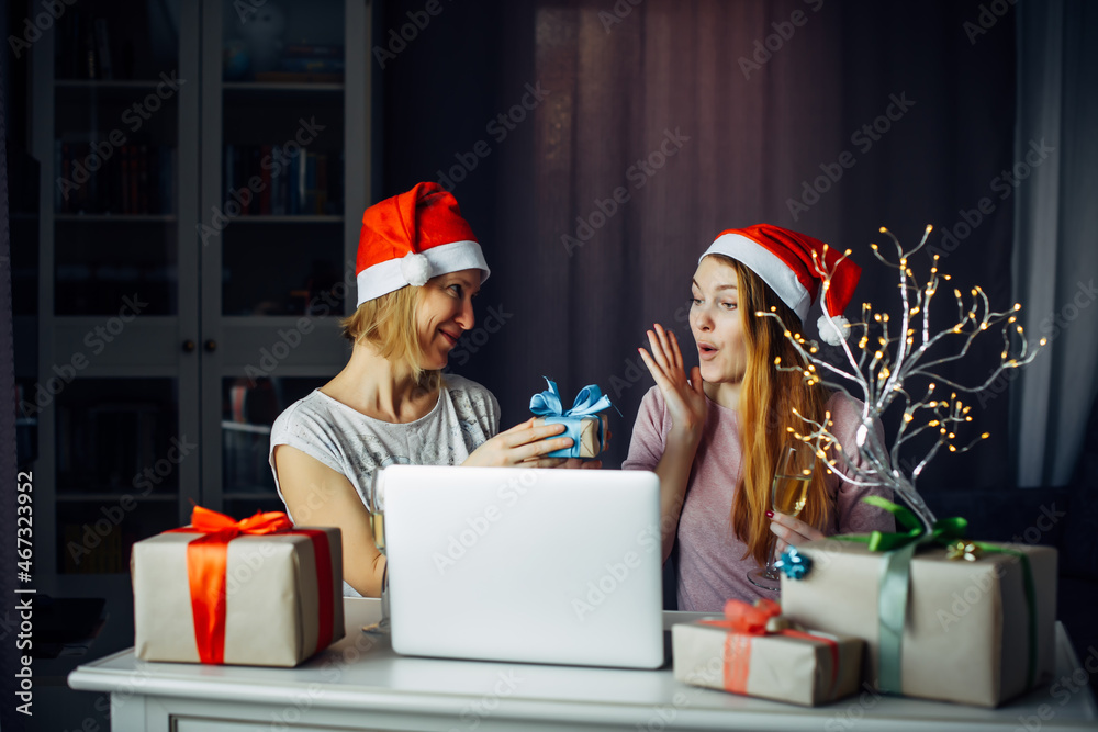 Fototapeta premium Two young women in Santa's hats exchange New Year's gifts in festive home interior. Joy of receiving a long-awaited gift for Christmas.