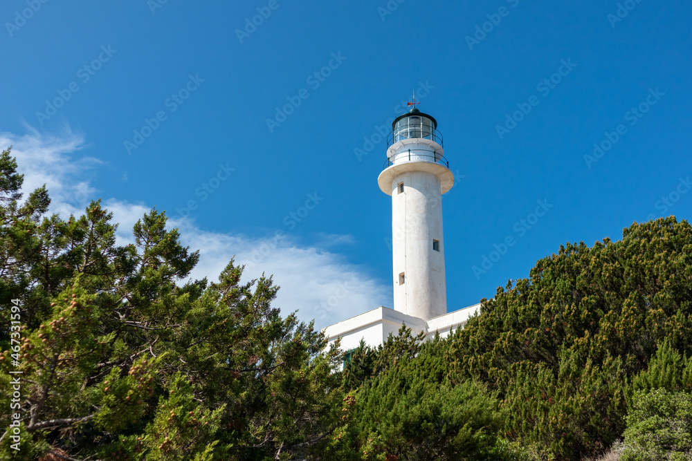 White lighthouse tall tower in pine bushes greenery on a bright clear blue sky with light clouds in Greece, Ionian sea. Scenic travel destination. Lefkada island