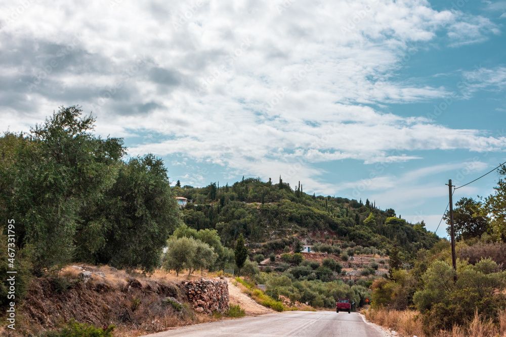 Fototapeta premium Scenic road to traditional Greek village with scenic cloudscape on green hills on Lefkada island, Greece. Travel Europe in summer by car