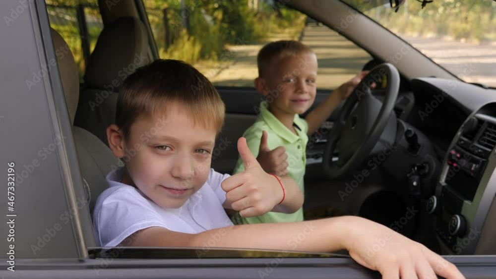 Two cheerful little brothers are sitting behind the wheel in a car, they are playing and giving a thumbs up.