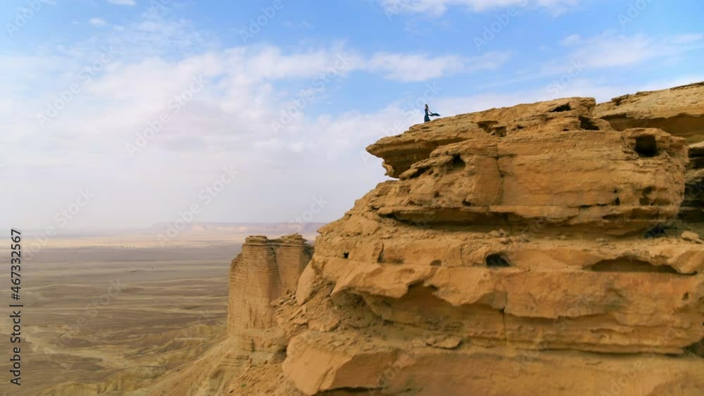 Aerial Panning Over A Woman Standing On The Edge Of A Soaring Cliff In An Arid Desert Under Hazy Skies - Arabian Desert, Saudi Arabia Aerial Panning Over A Woman Standing On The Edge Of A Soaring Cliff In An Arid Desert Under Hazy Skies - Arabian Desert, Saudi Arabia