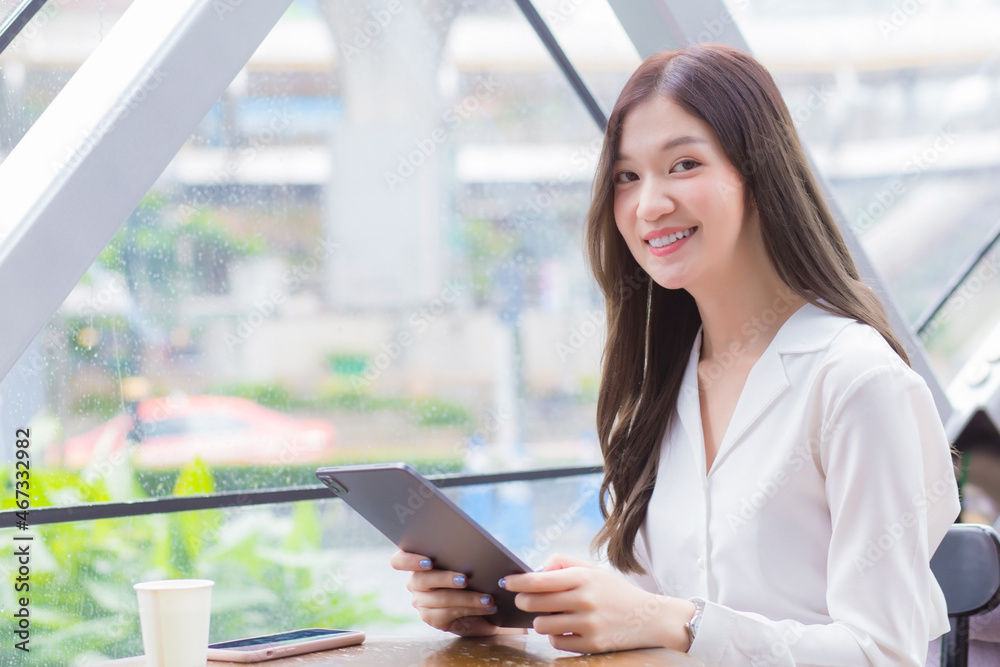 Young Asian business woman is looking at camera in her hands tablet or notepad while she is sitting at coffee shop among business background in the city. Young Asian business woman is looking at camera in her hands tablet or notepad while she is sitting at coffee shop among business background in the city.