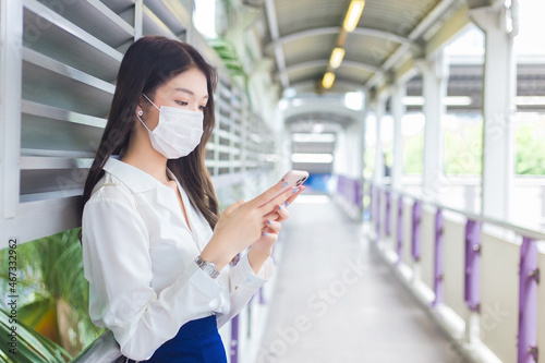 Young Asian beautiful business woman wearing a face mask stands on flyover of skytrain in town while uses her smartphone to send messages to office staff.