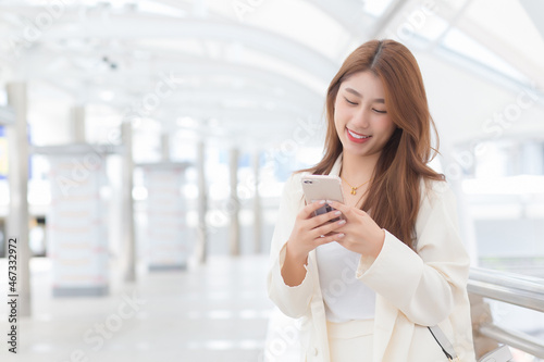Young Asian business woman in white suit smiles and holds a smartphone in her hands in happily workday at the outdoors city as a background.