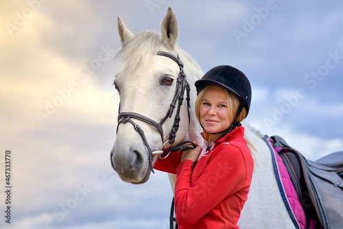 Portrait of a beautiful happy girl, young woman rider, equestrian smiling standing near white horse in helmet outdoors on sky background. Horseback riding sport. People love, care animals concept