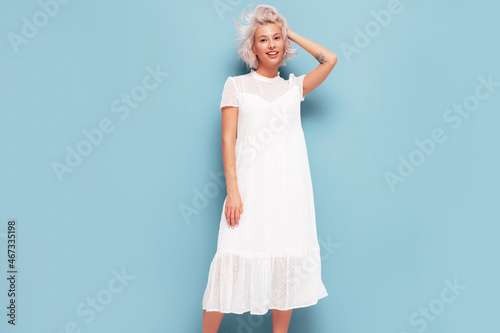 Portrait of young beautiful smiling female in trendy summer white dress. Sexy carefree blond woman posing near blue wall in studio. Positive model having fun indoors. Cheerful and happy