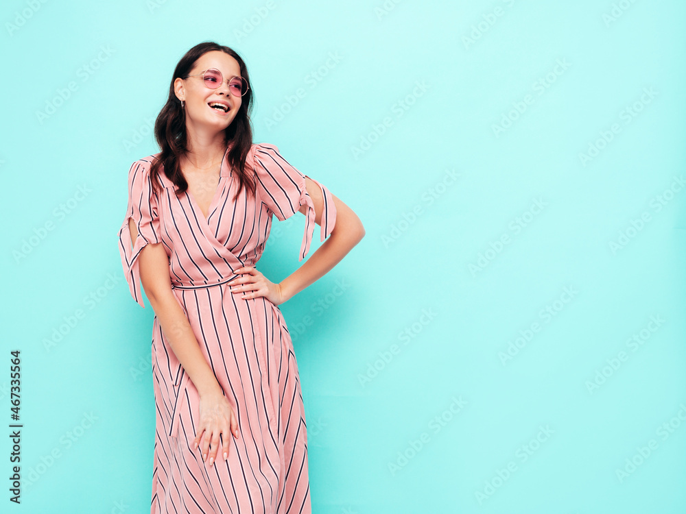 Portrait of young beautiful smiling female in trendy summer pink dress. Sexy carefree woman posing near blue wall in studio. Positive model having fun indoors. Cheerful and happy in sunglasses Portrait of young beautiful smiling female in trendy summer pink dress. Sexy carefree woman posing near blue wall in studio. Positive model having fun indoors. Cheerful and happy in sunglasses