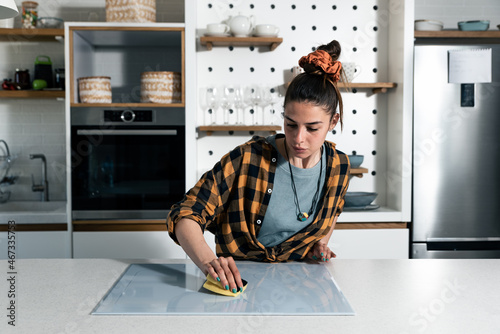 Young beautiful woman suffers from depression after a breakup or divorce and suffers from OCD cleans the kitchen cook top to calm her nerves. Serious stressed female rubbing the home with sponge. photo