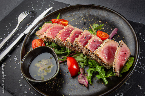 Close up of tuna slices withsesam seeds and fresh vegetables on a dark plate.