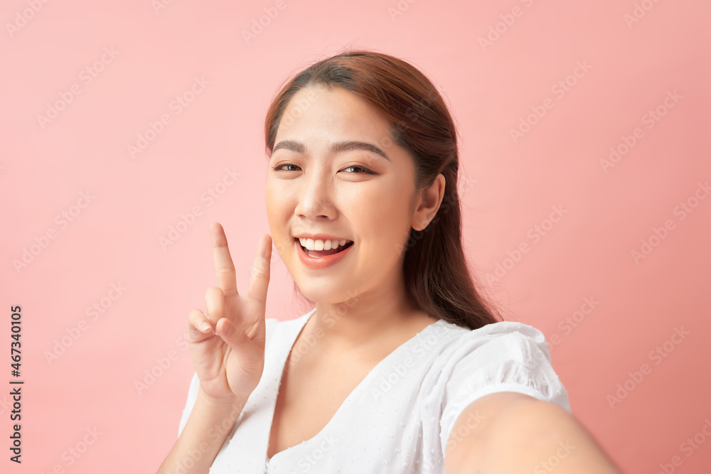 Young cheerful woman taking a self-portrait and gesturing v-sign against pink background Young cheerful woman taking a self-portrait and gesturing v-sign against pink background