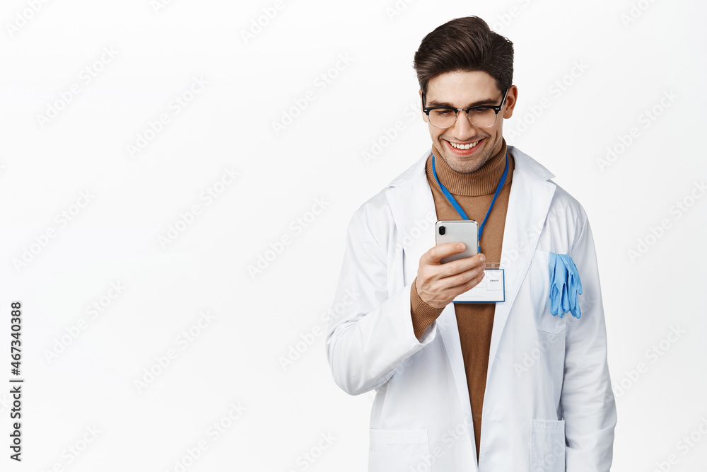 Smiling doctor using mobile phone, looking at screen, checking medical app, standing over white background in uniform Smiling doctor using mobile phone, looking at screen, checking medical app, standing over white background in uniform