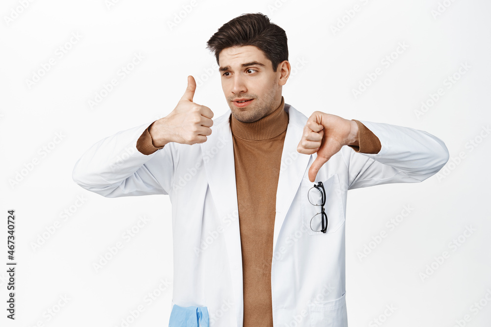 Indecisive healthcare worker showing thumb up and thumb down, pros or cons, making decision, standing in medical robe against white background Indecisive healthcare worker showing thumb up and thumb down, pros or cons, making decision, standing in medical robe against white background