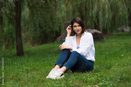 Attractive, happy and stylish brunette Caucasian girl posing while sitting on green grass in the park. Lifestyle and fashion concept. © Павел Костенко