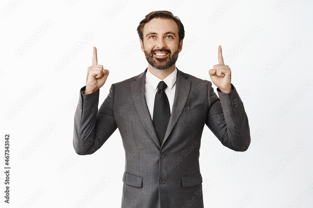 Happy businessman in suit pointing and looking up with pleased face, showing advertisement, standing over white background Happy businessman in suit pointing and looking up with pleased face, showing advertisement, standing over white background