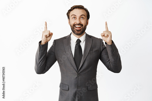Handsome businessman with beard, wearing suit, pointing fingers up and looking at advertisement, showing promo offer, white background