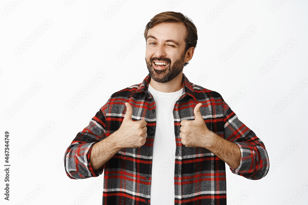 Enthusiastic male model winks and smiles, shows thumbs up, stands in checked shirt over white background