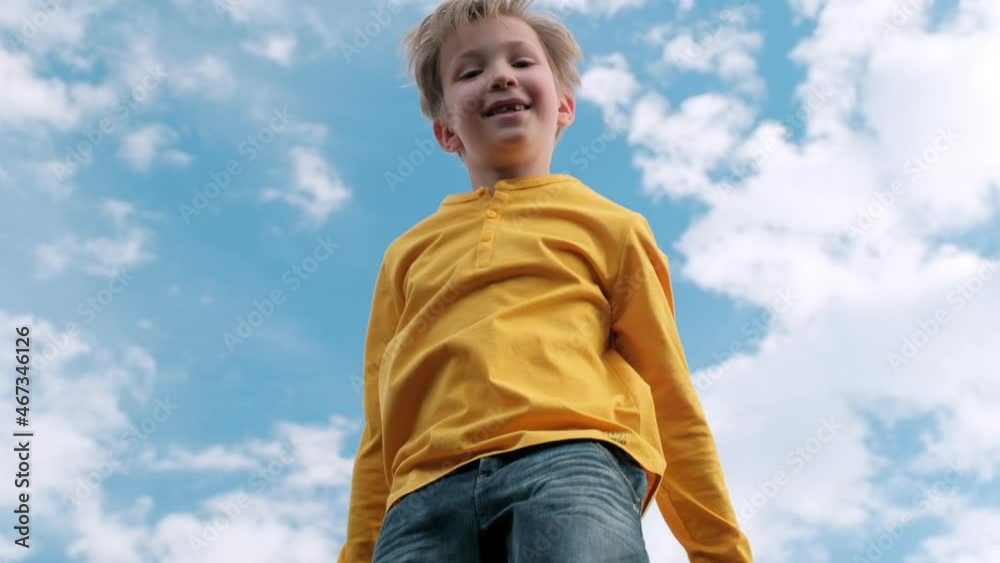 Happy child jumps very high on a trampoline against the blue sky. Blond boy jumps on a trampoline. Slow motion bottom view of a cheerful boy jumping on a trampoline. Children happiness.