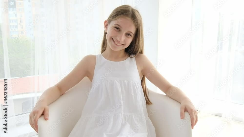 Beautiful teenage girl in white dress sitting, looking at the camera and smiling. Pretty female posing at home Beautiful teenage girl in white dress sitting, looking at the camera and smiling. Pretty female posing at home