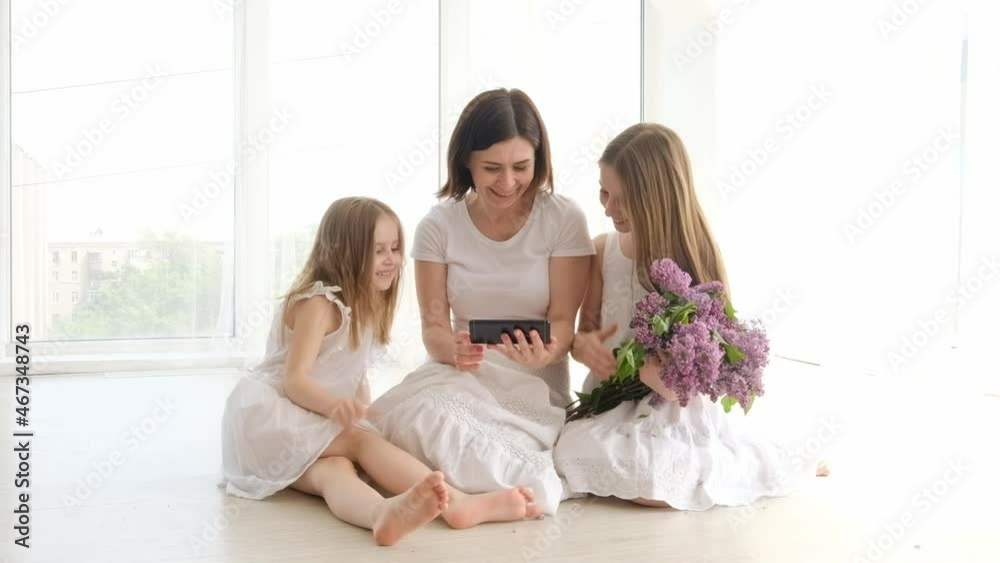 Mother with daughters in white dresses and with lilac bouquet looking photos on smartphone together. Children with parent and flowers with cell phone Mother with daughters in white dresses and with lilac bouquet looking photos on smartphone together. Children with parent and flowers with cell phone