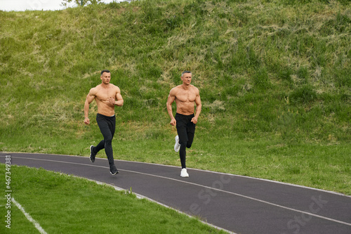 Handsome muscular guys with naked torso spending free time for running on fresh air. Active exercises of two bodybuilders. Sport activity on stadium. photo