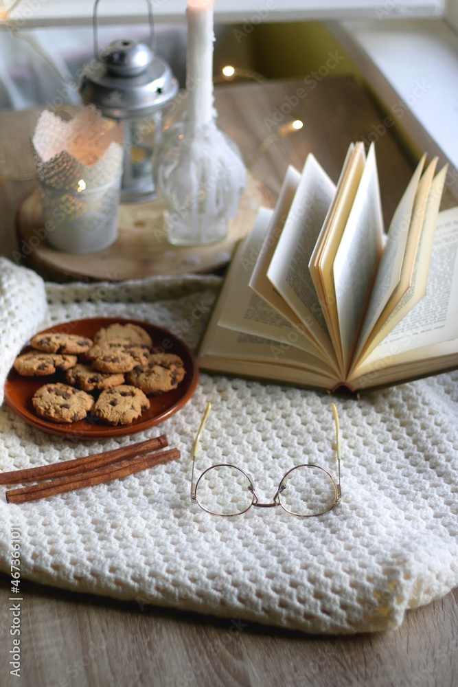 Plate of chocolate chip cookies, cinnamon sticks, soft blanket, open book and lit candles. Hygge at home. Selective focus. Plate of chocolate chip cookies, cinnamon sticks, soft blanket, open book and lit candles. Hygge at home. Selective focus.