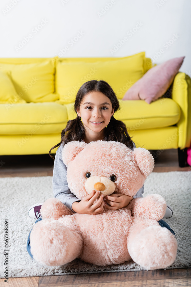 cheerful preteen girl holding soft toy while sitting on carpet at home.