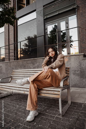  Tall stylish girl with oversized jacket sitting on the bench of the city background