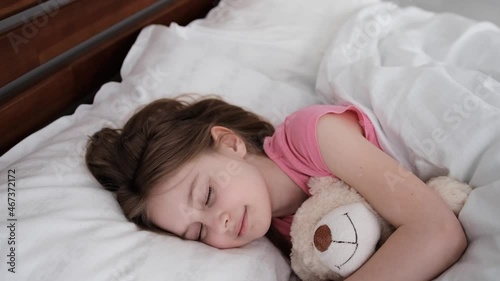 Little beautiful girl lying in the bed, hugging teddy bear and resting with smali on her face. Cute preteen child sleeping with toy. Female kid napping in the bedroom photo