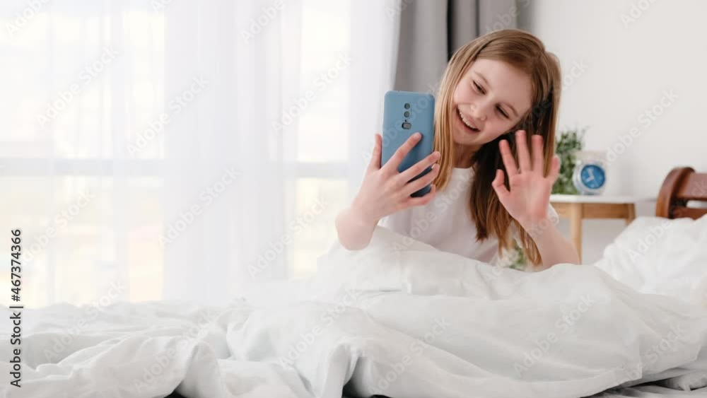Beautiful preteen little girl sitting in the bed and talking by video call on smartphone. Cute smiling kid staying in the bedroom with cell phone. Child and modern technologies Beautiful preteen little girl sitting in the bed and talking by video call on smartphone. Cute smiling kid staying in the bedroom with cell phone. Child and modern technologies