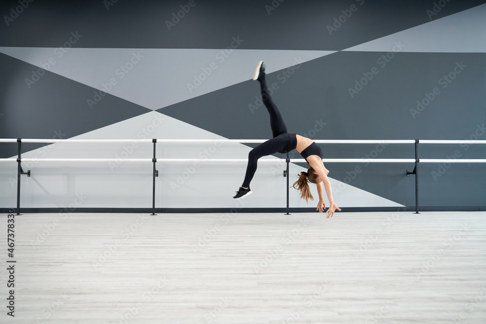 Side view of strong fit woman practicing wheel in jump, doing split in air. Attractive flexible sportswoman training in dance hall with handrails. Concept of choreography, healthy lifestyle.