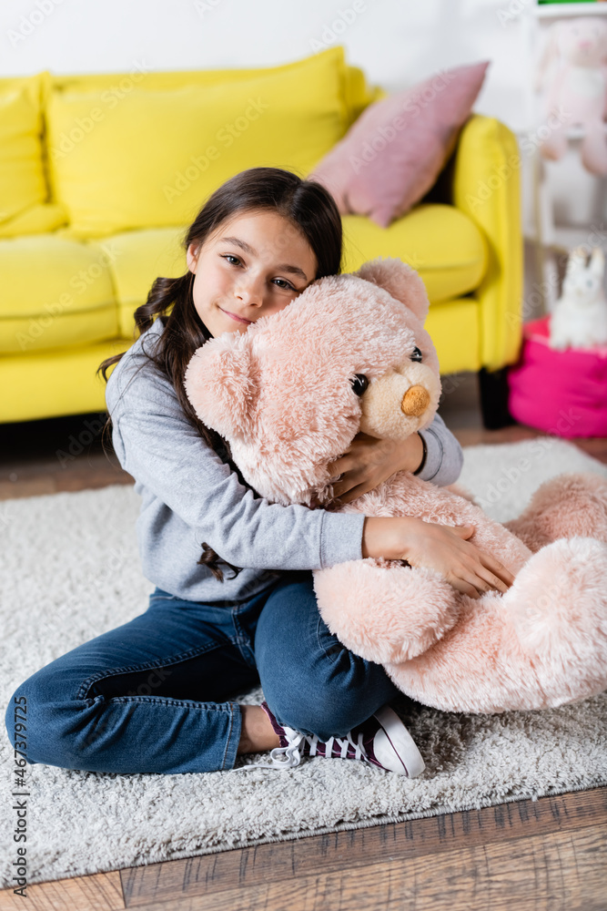 pleased preteen girl hugging soft toy while sitting on carpet at home ...