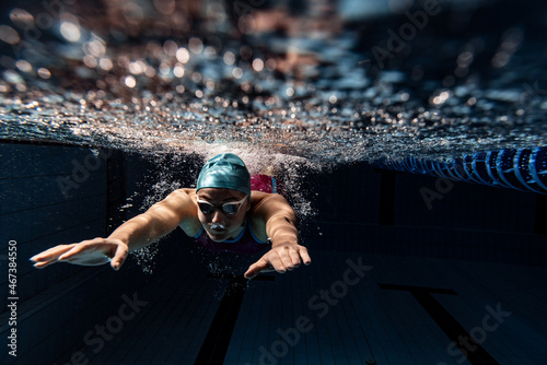 Underwater view of swimming movements details. One female swimmer in swimming cap and goggles training at pool, indoors.