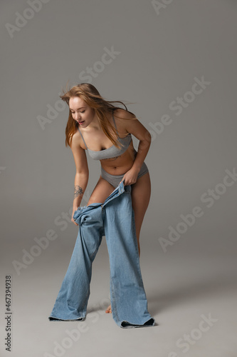 Young beautiful red-headed woman putting blue jeans jumpersuit on isolated over gray studio background. Weight Loss concept.