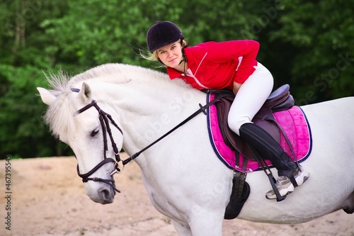 Portrait of a beautiful girl, young woman rider, equestrian on white horse in helmet and polo shirt, riding outdoors, galloping, looking at camera. Horseback riding sport.