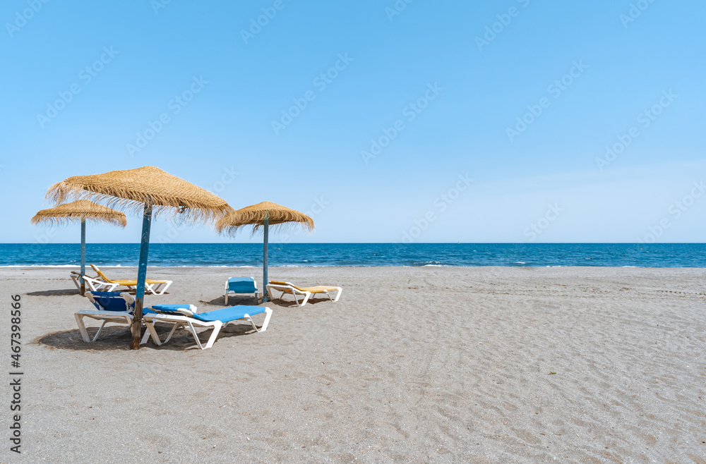 Sombrillas de madera y paja y hamacas sobre la arena de la playa y las aguas del mar mediterráneo un día soleado con cielo azul. Desde Málaga, Costa del sol, Andalucía, España.