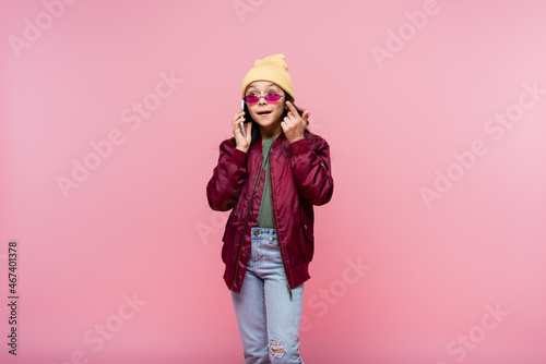 preteen girl in stylish outfit and sunglasses talking on smartphone isolated on pink.
