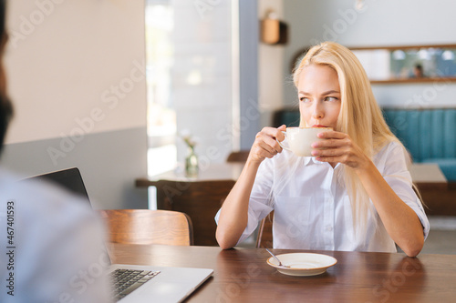 Front view of happy attractive blonde woman sitting in front of unrecognizable young man at table of cafe and drinking hot coffee, looking away. Romantic date in cafe, couple enjoying time.