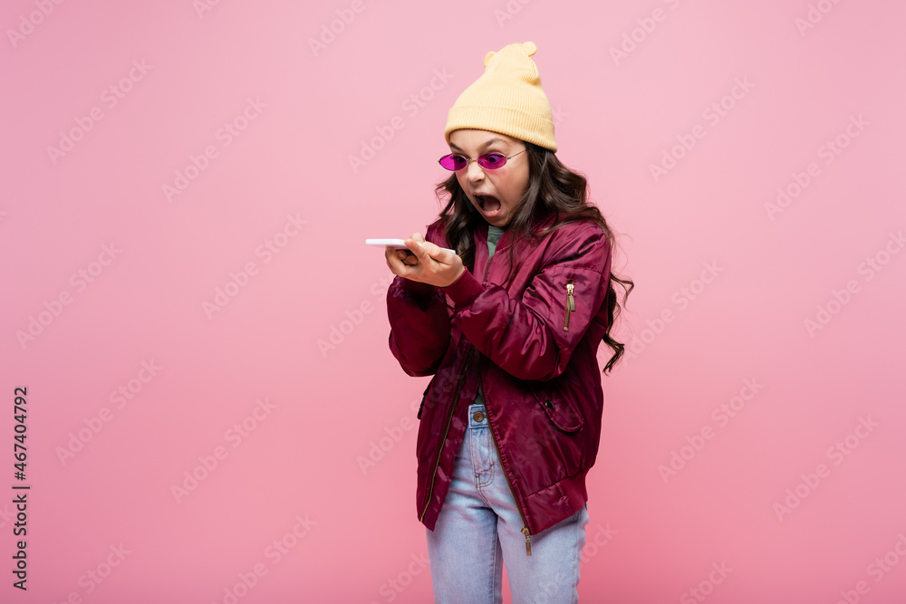 angry preteen girl in stylish outfit and sunglasses looking at smartphone and screaming isolated on pink. angry preteen girl in stylish outfit and sunglasses looking at smartphone and screaming isolated on pink.